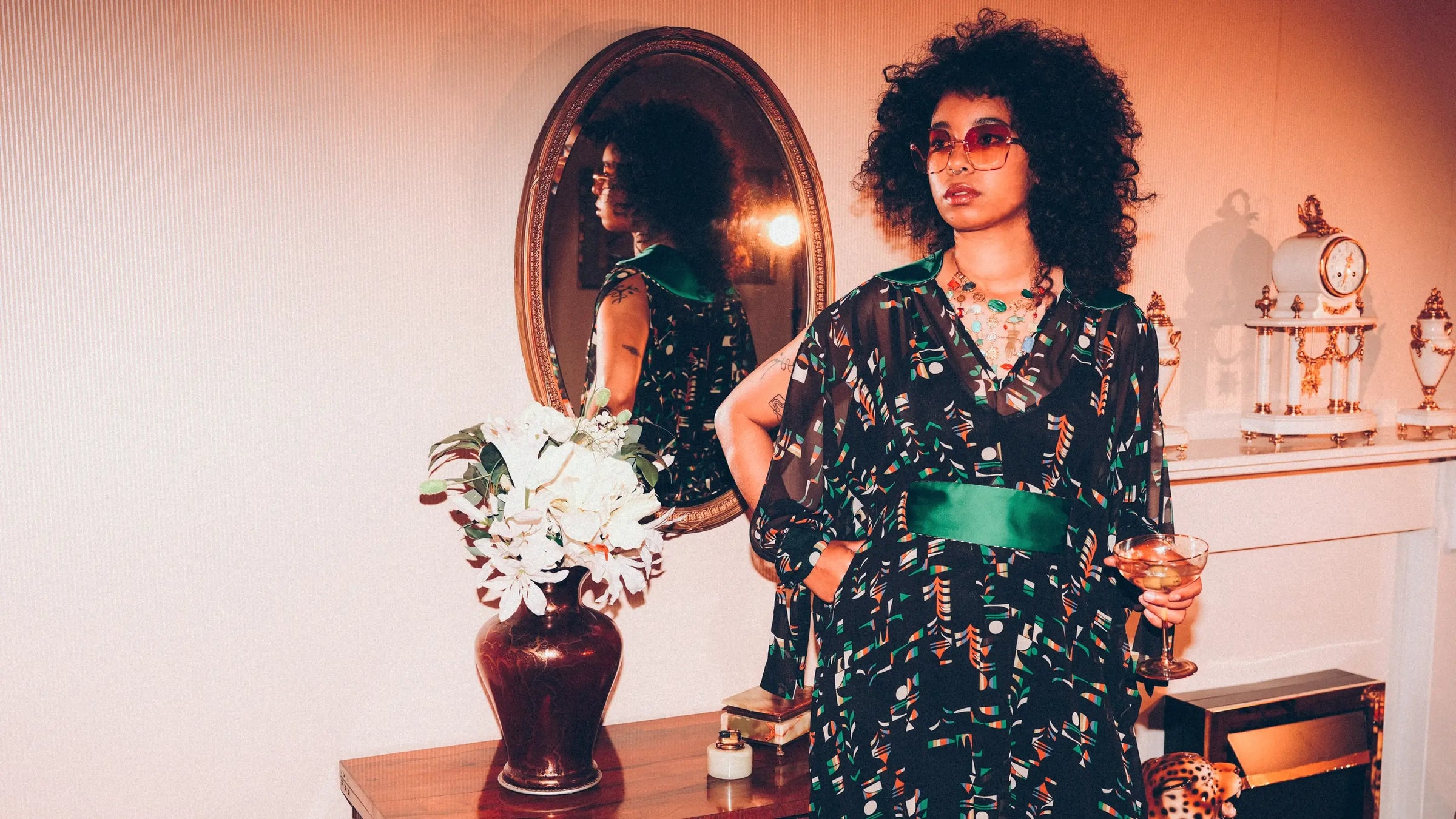Yazz wears Holly Campbell Studio's 'Lady Jo jo maxi Kaftan. Woman with blow-out afro in a patterned dress standing in front of a mirror with decorative items on a shelf.