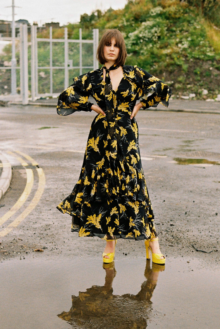 Model wears the Angie Dress Beautiful Oak leaf black and yellow chiffon print maxi dress with spiral frill sleeves and cascading frill at the lower part of the skirt. She stands in a puddle wearing bright yellow high heels in an urban setting.