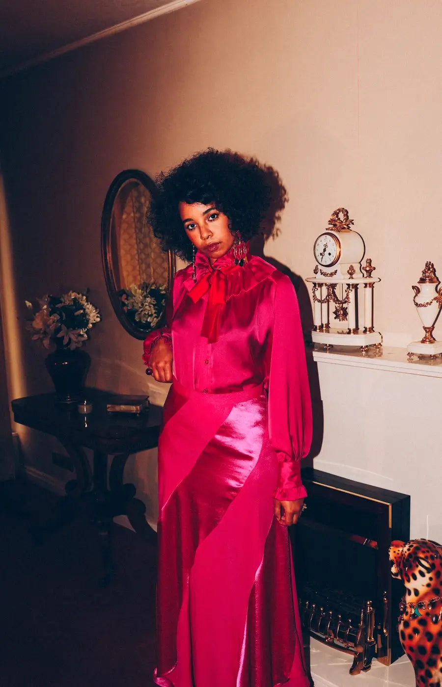 Yazz wears the Estelle satin shirt in bright pink by Holly Campbell studio. Woman in a pink dress standing in a room with decorative items.