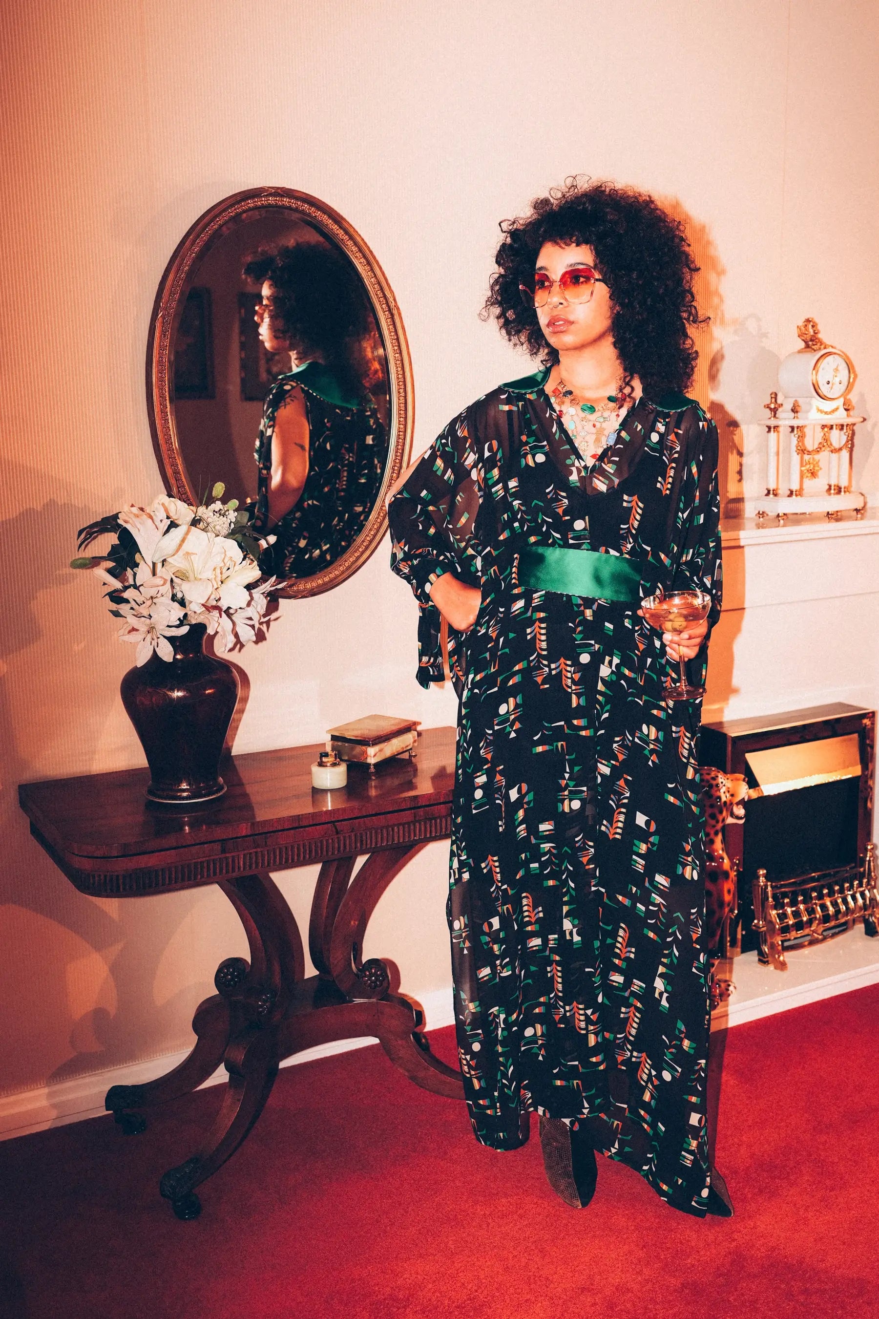 Edinburgh Musician Yazz, models the patterned Madame Jojo kaftan by Edinburgh fashion designer 
Holly Campbell. She is standing in a vintage-style living room with a mirror and a table.