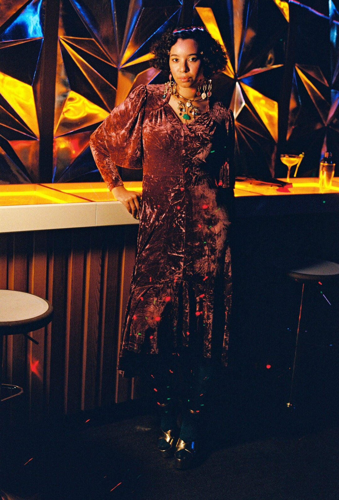 Model leaning on a bar in a dusky rose crushed velvet midi dress with cape sleeves and a high-low hem